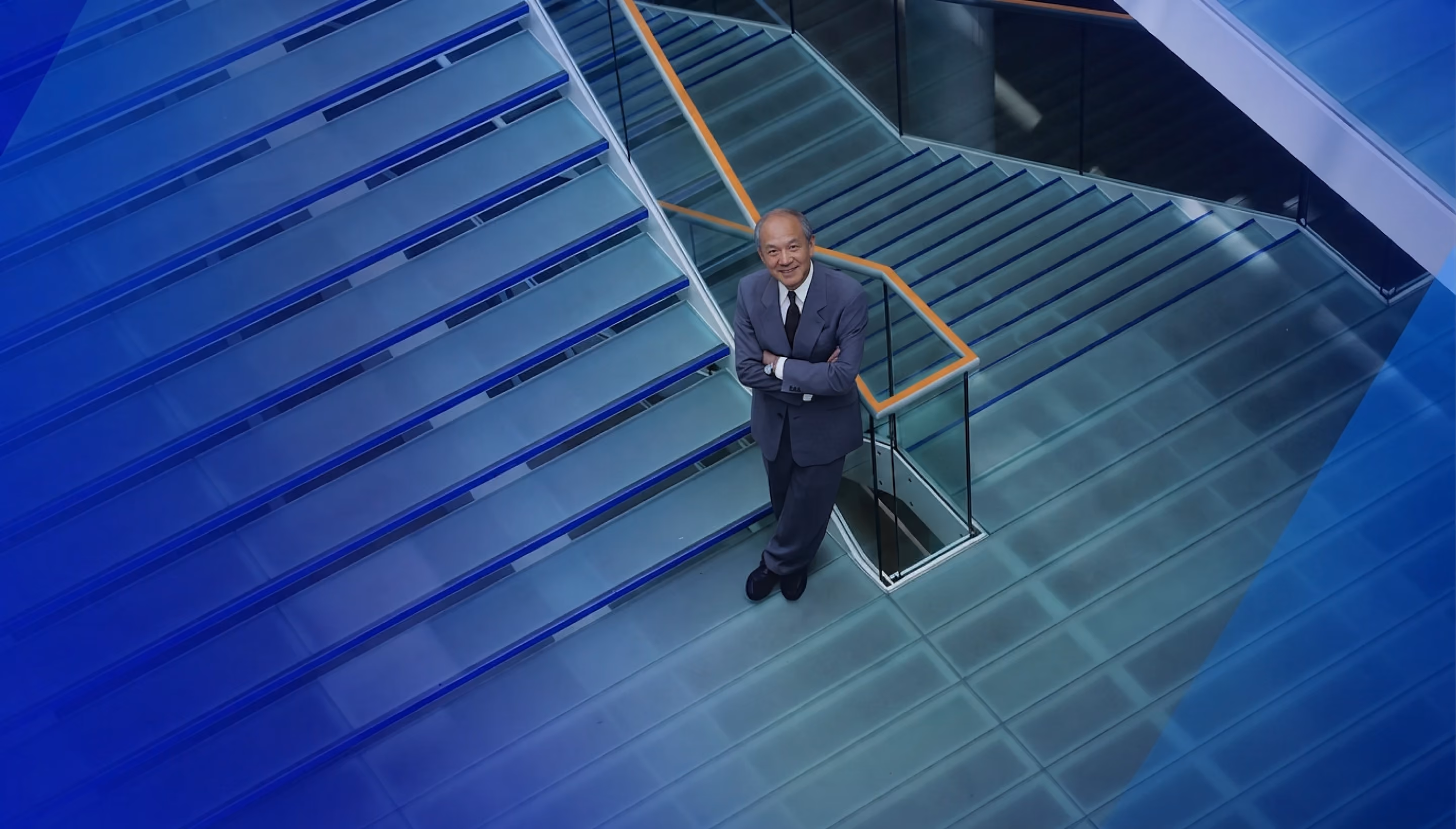Man in suit standing on glass staircase with hands crossed, smiling and looking up.