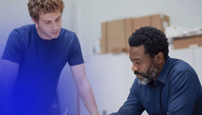 Two men in a workspace, one standing and one sitting, focused on work or discussion.