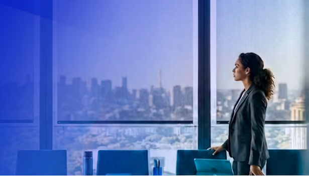 Businesswoman in suit standing in office looking out large window at city skyline.