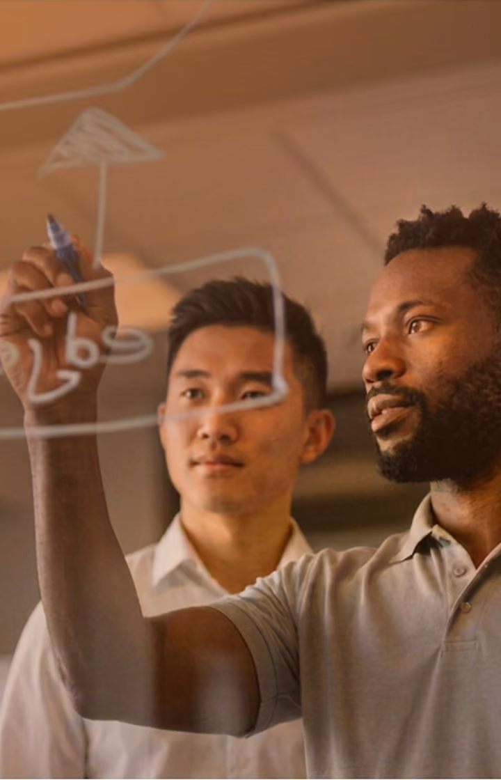 Two men collaborating while one draws a flowchart on a transparent board with a marker.