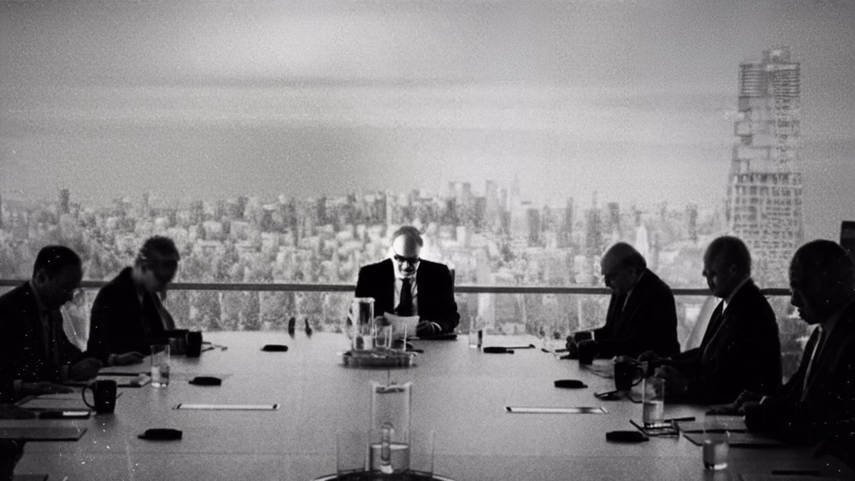 Black-and-white photo of six people in suits sitting around a long conference table in a high-rise room.