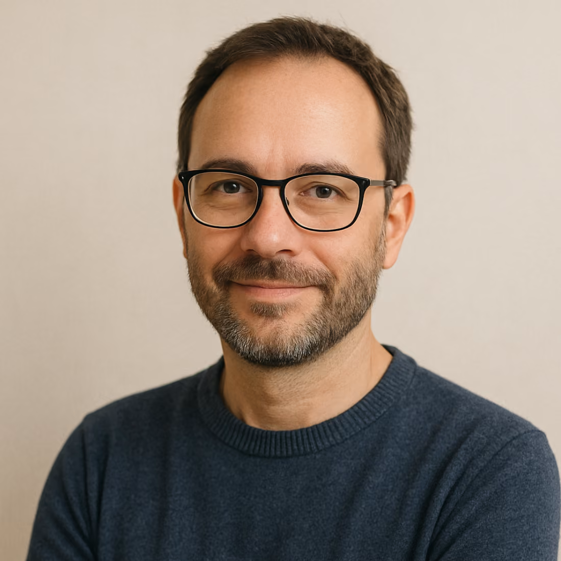 Homme aux cheveux bruns courts et barbe courte, portant des lunettes noires et un pull bleu, souriant légèrement devant un fond clair.