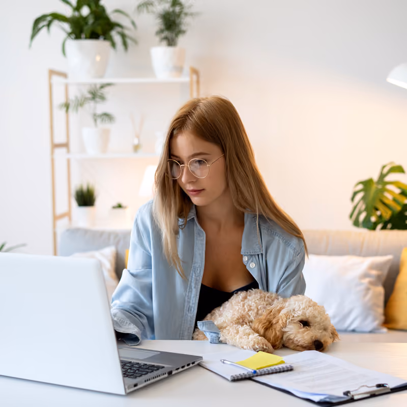 Jeune femme aux cheveux longs et lunettes regardant un ordinateur portable blanc, avec un petit chien bouclé allongé sur la table devant elle, dans un salon lumineux avec des plantes en arrière-plan.