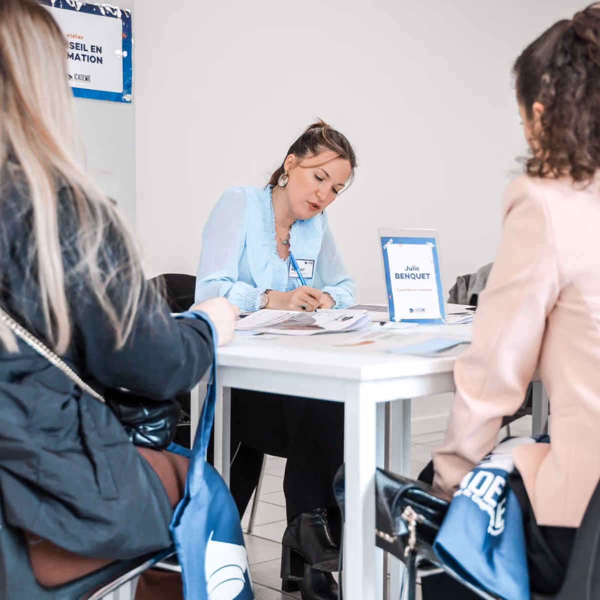 Une femme en chemisier bleu clair écrit sur des documents à une table avec un panneau indiquant « Julie BENQUET » tandis que deux autres femmes sont assises face à elle dans une réunion ou un entretien dans une pièce claire.