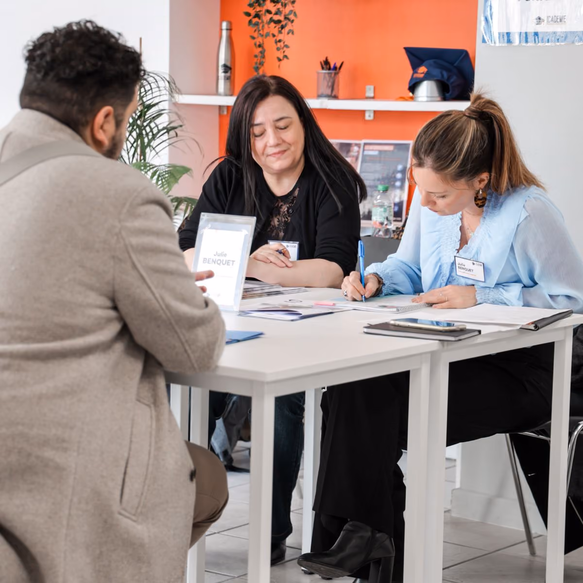 Trois personnes assises autour d'une table blanche dans un bureau; une femme au centre sourit, une femme à droite écrit dans un carnet, un homme de dos écoute.