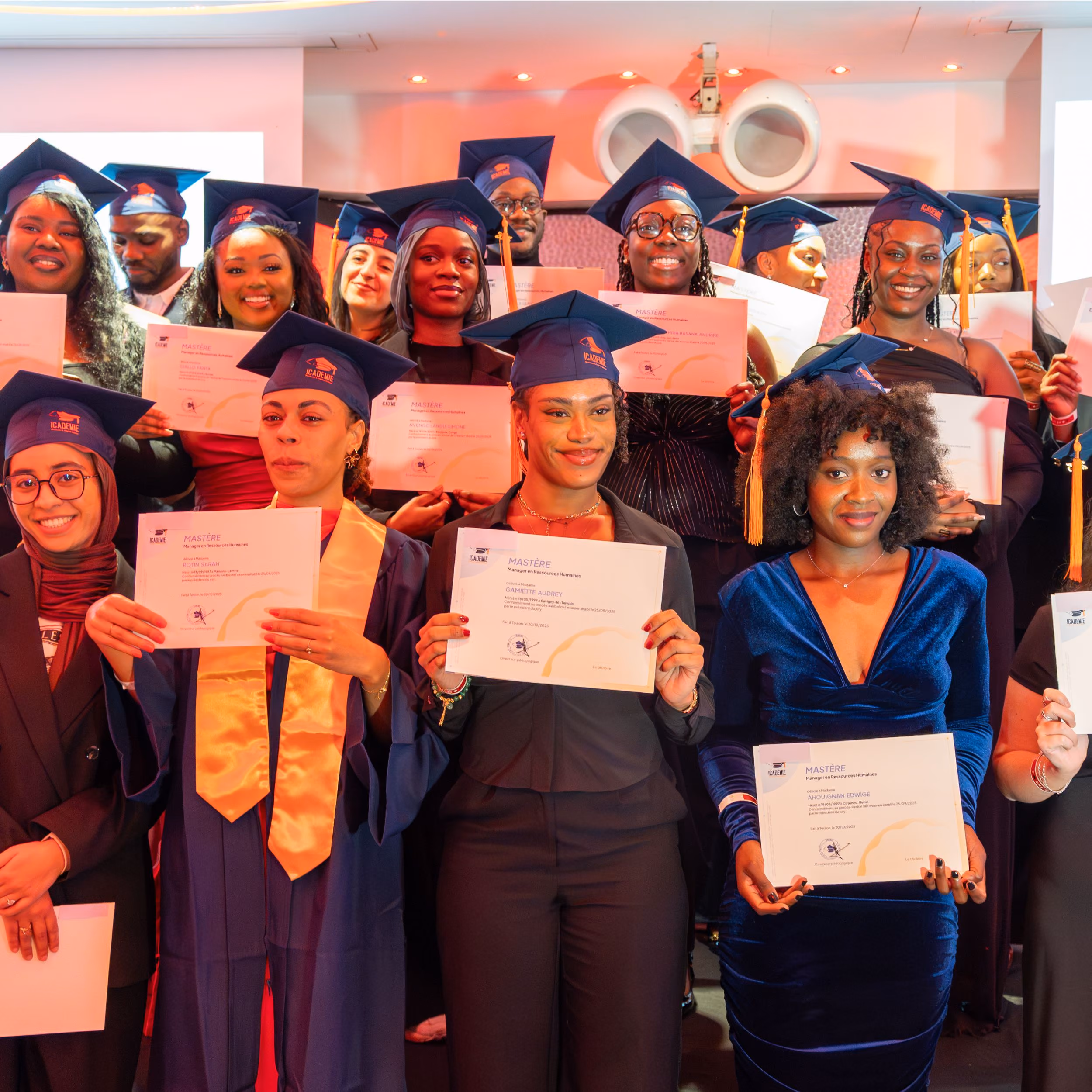 Un groupe de diplômés souriants portant des toques et des robes de cérémonie tenant leurs diplômes, posant pour une photo de groupe lors d'une cérémonie de remise de diplômes en intérieur.
