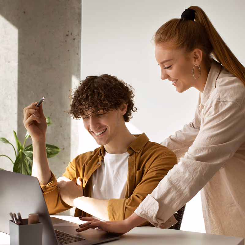 Un homme souriant assis à une table devant un ordinateur portable avec un stylo en main, une femme souriante se tenant à côté de lui en tapant sur le clavier.