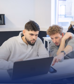 Deux hommes assis à une table regardant un ordinateur portable, l'un d'eux pointant l'écran en expliquant quelque chose.