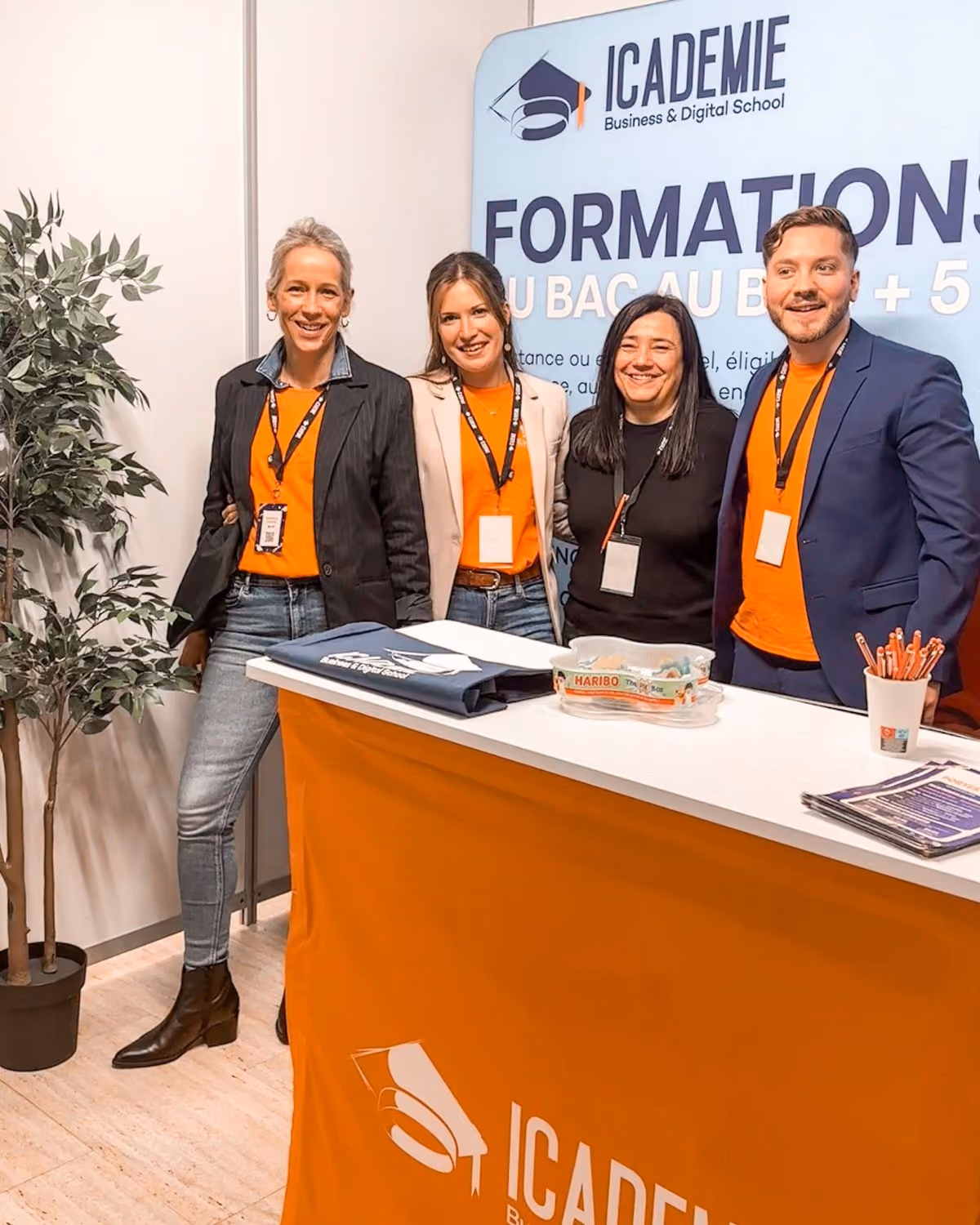 Quatre personnes souriantes, trois femmes et un homme, posent derrière un stand avec une nappe orange marquée du logo ICADÉMIE, une école de commerce et numérique, dans un environnement intérieur avec une plante à gauche et une affiche promotionnelle en arrière-plan.