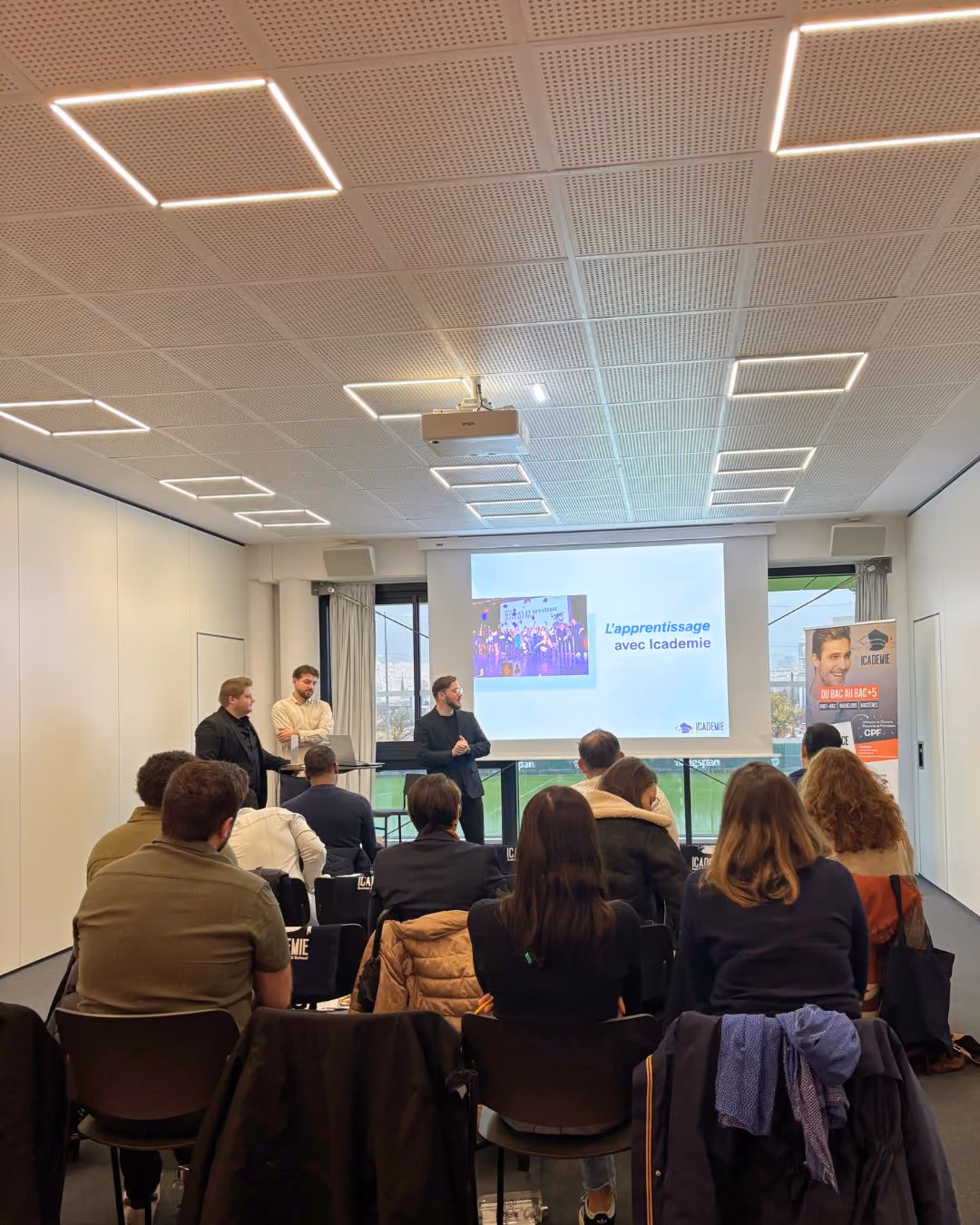 Une salle de classe avec plusieurs participants assis de dos face à trois présentateurs debout devant un grand écran affichant une diapositive intitulée « L'apprentissage avec l'académie ». La salle est lumineuse avec un plafond équipé de lumières carrées et des murs blancs. Une bannière est visible à droite de l'écran. Les participants semblent attentifs à la présentation.