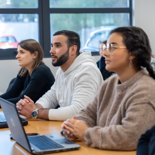 Quatre jeunes adultes assis à une table avec des ordinateurs portables, écoutant attentivement lors d'une réunion ou d'une classe dans une salle lumineuse avec de grandes fenêtres.