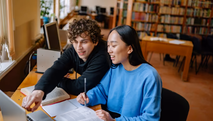 Deux étudiants, un homme aux cheveux bouclés en sweat noir et une femme aux cheveux longs noirs en sweat bleu, travaillent ensemble sur un ordinateur portable dans une bibliothèque publique avec des étagères pleines de livres en arrière-plan.