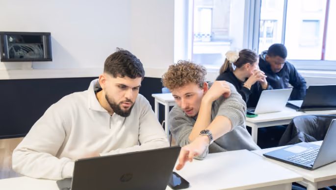 Deux jeunes hommes collaborent devant un ordinateur portable dans une salle de classe moderne avec deux autres étudiants travaillant en arrière-plan.