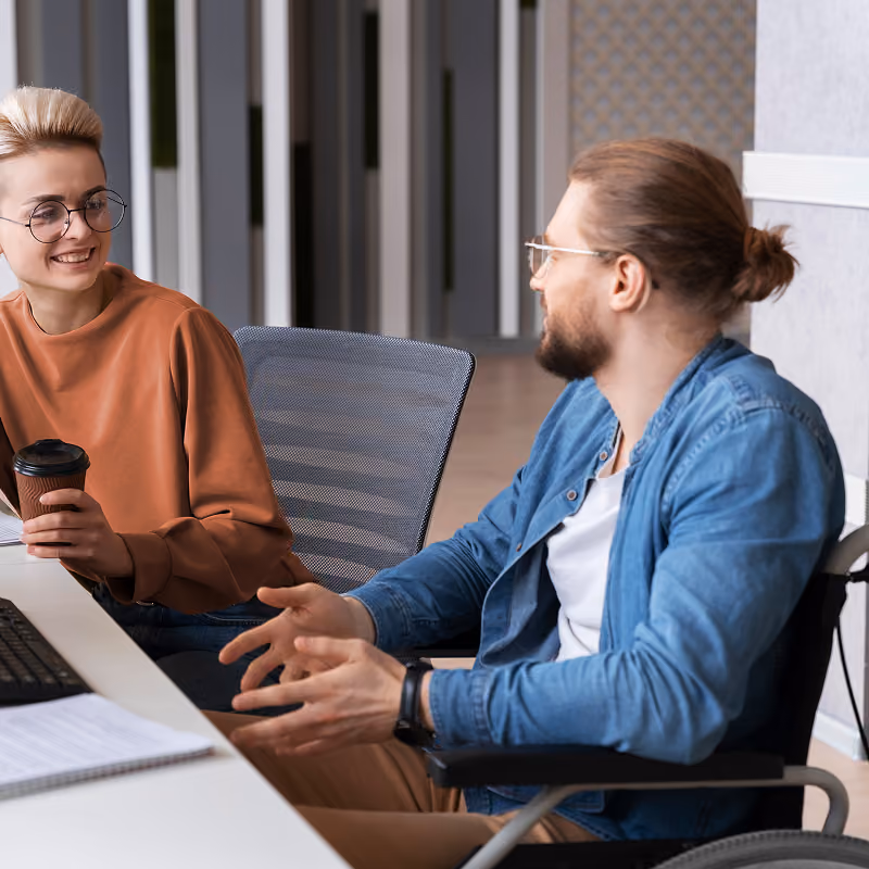 Deux collègues discutent dans un bureau moderne; une femme avec des lunettes tient un café et sourit à un homme avec lunettes et barbe qui gesticule.
