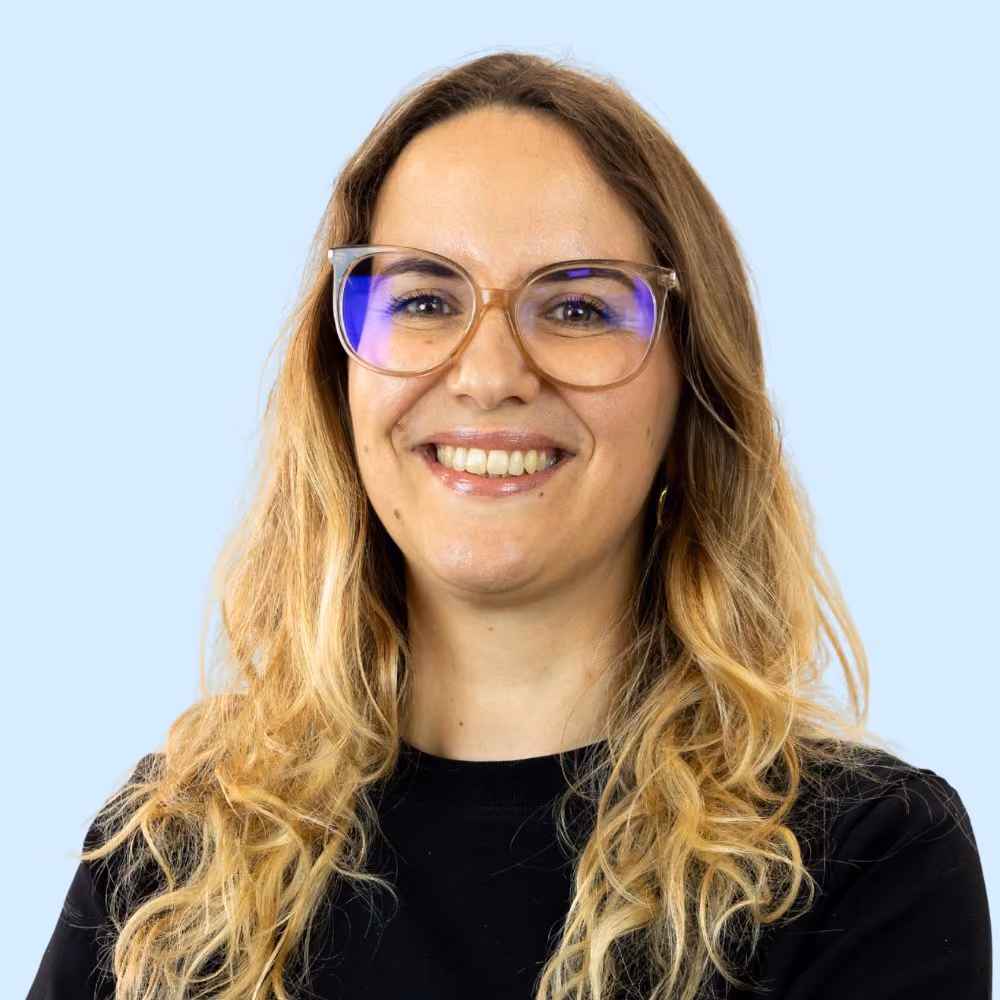 Femme souriante aux cheveux longs blonds portant des lunettes transparentes et un pull noir, sur fond bleu clair.
