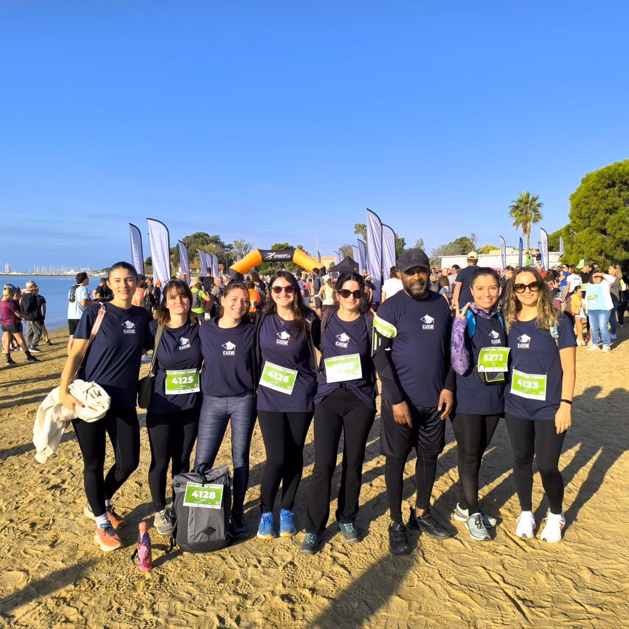 Groupe de huit coureurs portant des maillots bleu marine unis formant une ligne côte à côte sur une plage au lever du soleil, avec des dossards verts et souriant vers la caméra, avec un public et des drapeaux blancs en arrière-plan sous un ciel bleu clair.
