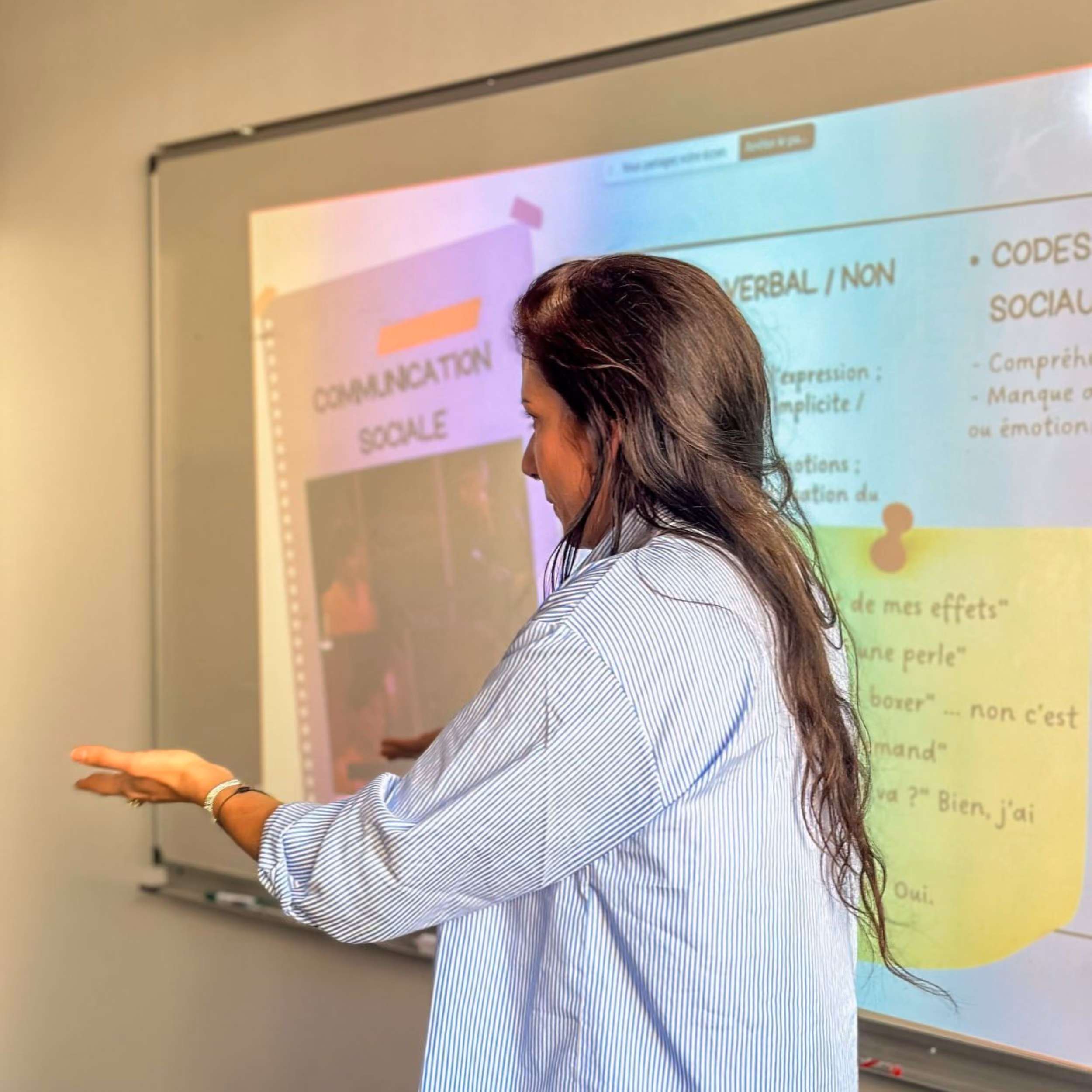 Une femme en chemise rayée donnant une présentation devant un tableau blanc avec diapositives projetées sur la communication sociale.