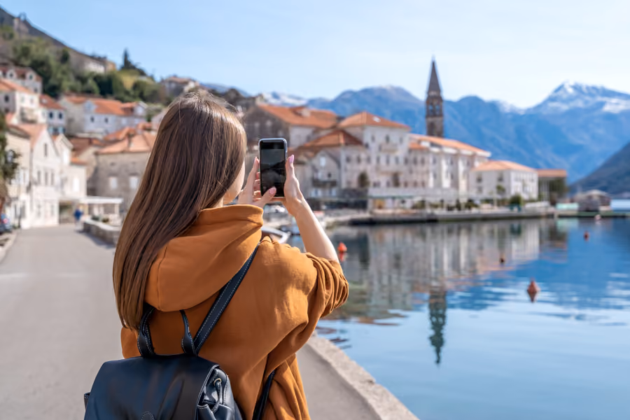 Une femme aux cheveux longs portant un sweat à capuche marron et un sac à dos noir prend une photo avec son téléphone sur une promenade près de l'eau, avec des bâtiments anciens à toits orange et des montagnes enneigées à l'arrière-plan.