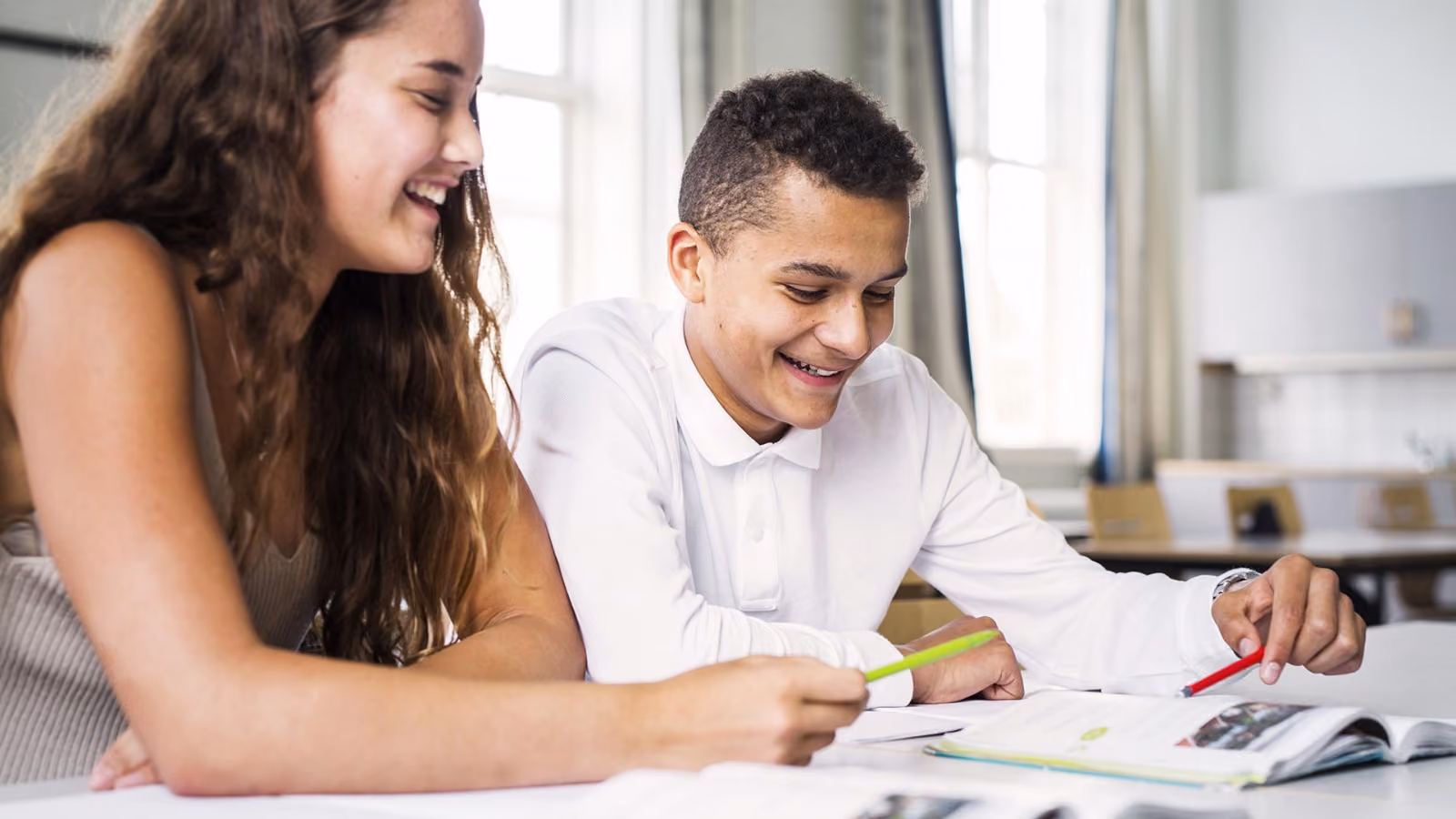 Deux adolescents souriants, un garçon et une fille, travaillent ensemble en classe, regardant un livre ouvert et tenant des crayons colorés.