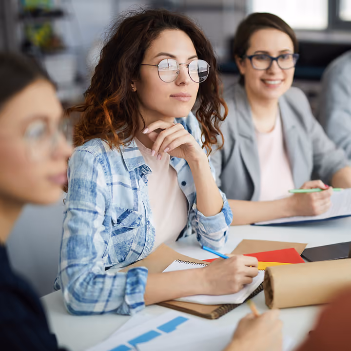 Jeunes femmes assises à une table lors d'une réunion ou d'un atelier, certaines prennent des notes sur des cahiers, l'ambiance est professionnelle et collaborative.