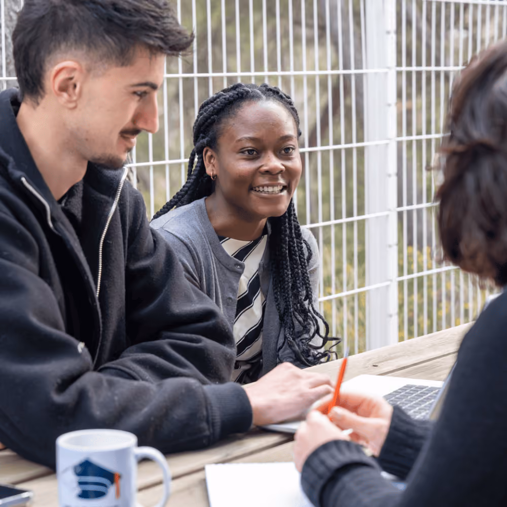 Trois jeunes adultes assis autour d'une table en bois à l'extérieur, discutant avec un ordinateur portable ouvert, une tasse blanche et un smartphone posés sur la table.