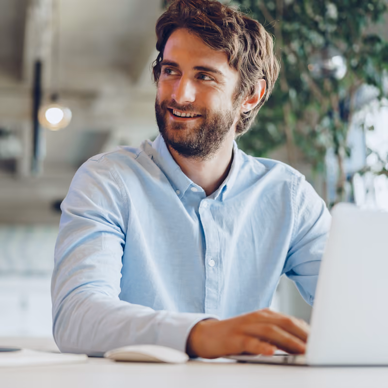 Un homme souriant portant une chemise bleue, assis à une table et utilisant un ordinateur portable dans un bureau moderne avec une plante en arrière-plan.