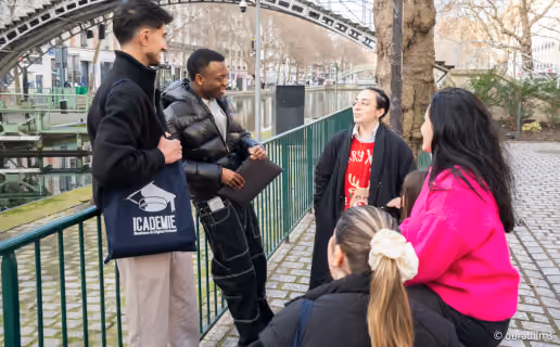 Un groupe de cinq jeunes discutant près d'un canal urbain, avec un pont métallique en arrière-plan et une clôture verte le long du chemin pavé.