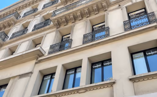 Façade d'un immeuble haussmannien beige avec plusieurs balcons en fer forgé et fenêtres à volets noirs reflétant un ciel bleu.