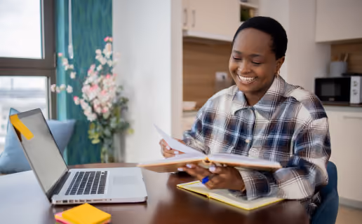 Une femme souriante assise à une table avec un ordinateur portable ouvert, tenant un dossier et feuilletant des documents dans une cuisine lumineuse.