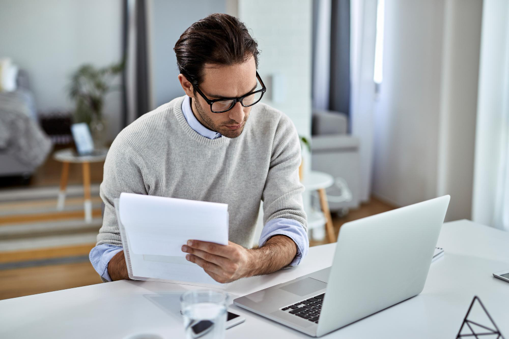 Homme à lunettes lisant une fiche devant son ordinateur portable