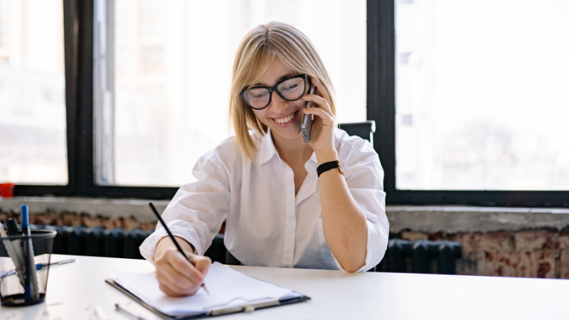 Femme blonde à lunettes souriante au téléphone, prenant des notes à son bureau