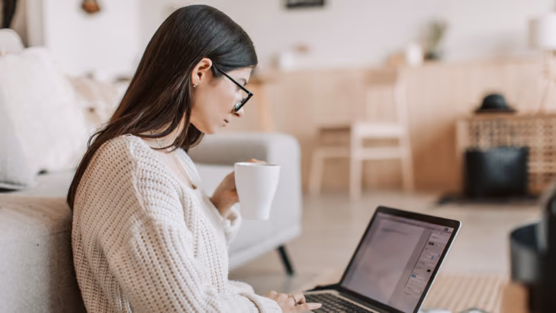 Jeune femme à lunettes travaillant sur un ordinateur portable, une tasse à la main, dans son salon