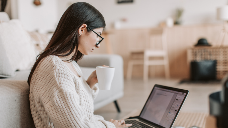 Jeune femme à lunettes travaillant sur un ordinateur portable, une tasse à la main, dans son salon
