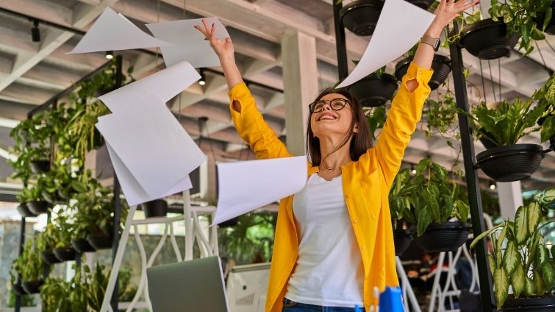 Femme en veste jaune lançant joyeusement des feuilles de papier en l'air dans un bureau