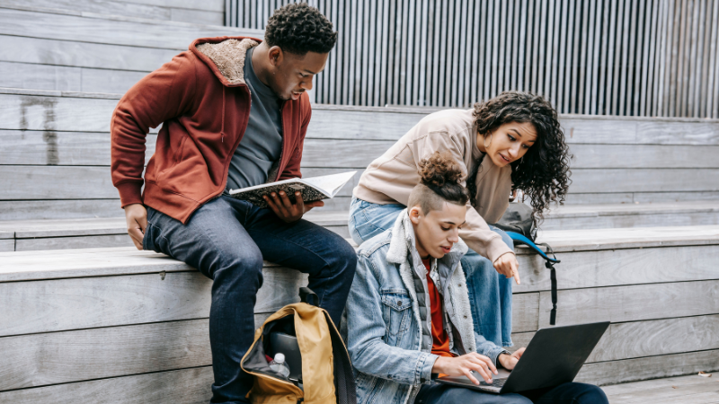 Trois étudiants assis sur des gradins en bois, révisant avec un livre et un ordinateur portable