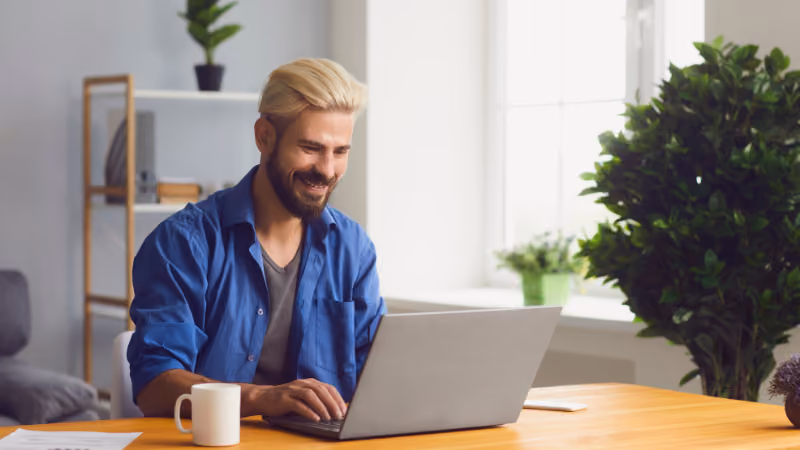 Homme barbu souriant en chemise bleue travaillant sur un ordinateur portable
