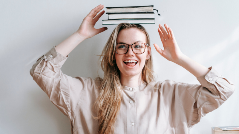 Jeune femme à lunettes riant avec une pile de livres en équilibre sur la tête