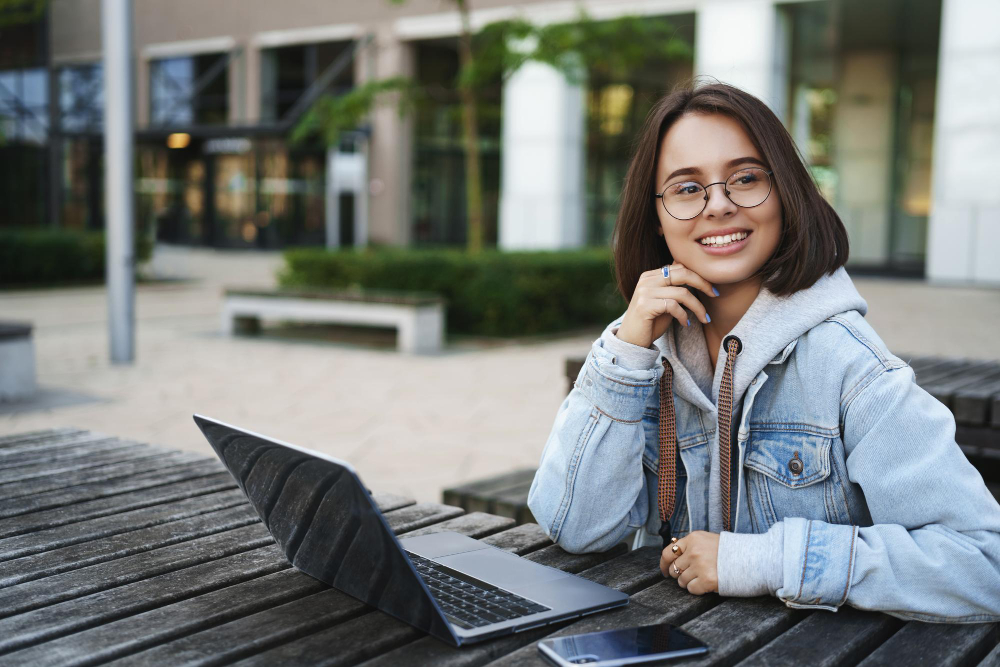 Étudiante souriante à lunettes assise à une table en extérieur avec son ordinateur portable