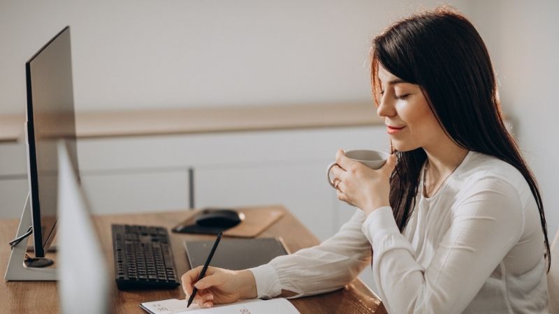 Femme prenant des notes une tasse à la main devant son ordinateur