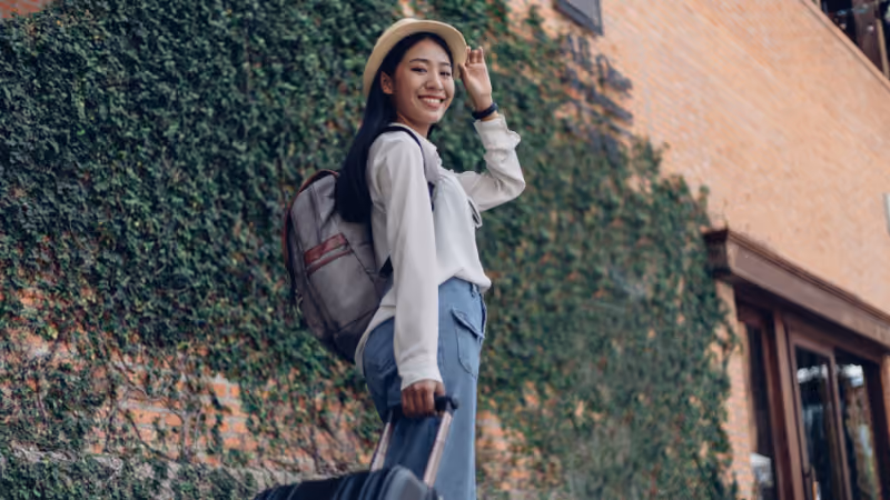 Étudiante souriante avec un chapeau et un sac à dos devant un mur couvert de lierre