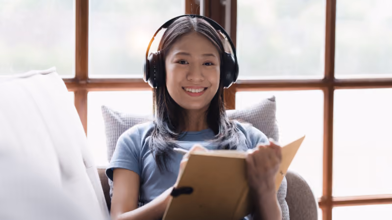 Jeune femme souriante avec un casque audio lisant un livre sur un canapé