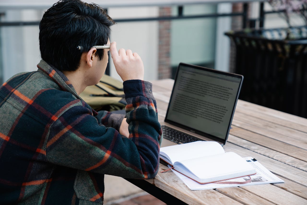 Jeune homme ajustant ses lunettes devant son ordinateur portable et un cahier ouvert