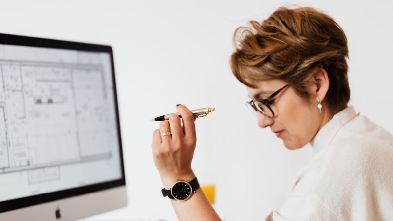 Femme à lunettes pensive, stylo à la main, devant un écran affichant un plan