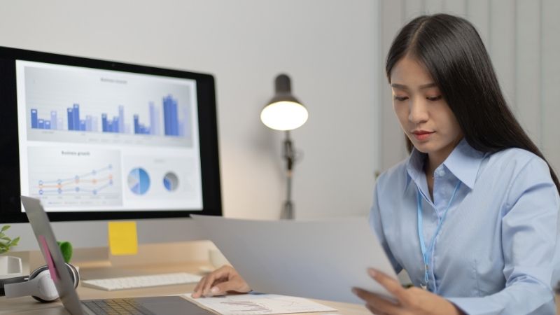 Jeune femme examinant un document devant un écran affichant des graphiques