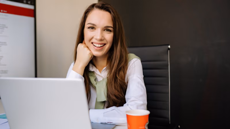 Jeune femme souriante accoudée à son bureau devant un ordinateur portable