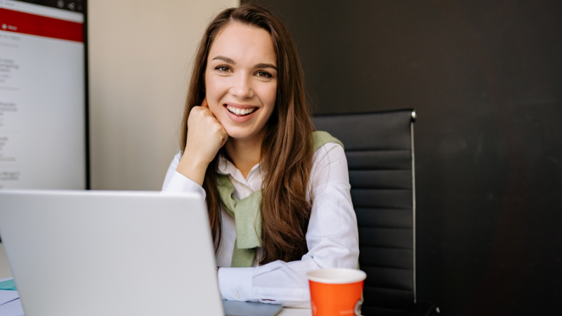 Jeune femme souriante accoudée à son bureau devant un ordinateur portable