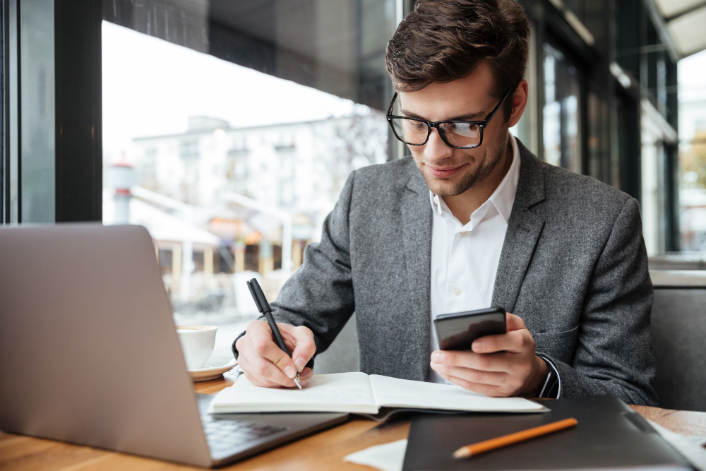 Homme à lunettes en veste prenant des notes en consultant son téléphone