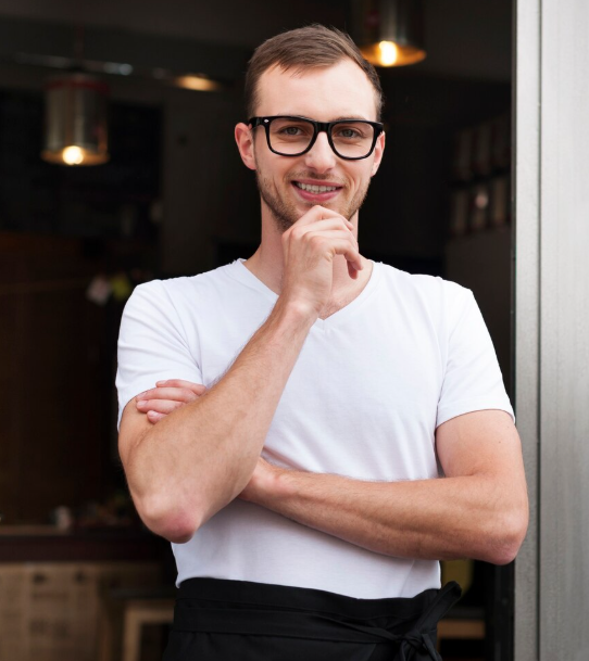 Restaurateur souriant en t-shirt blanc et tablier à l'entrée de son établissement