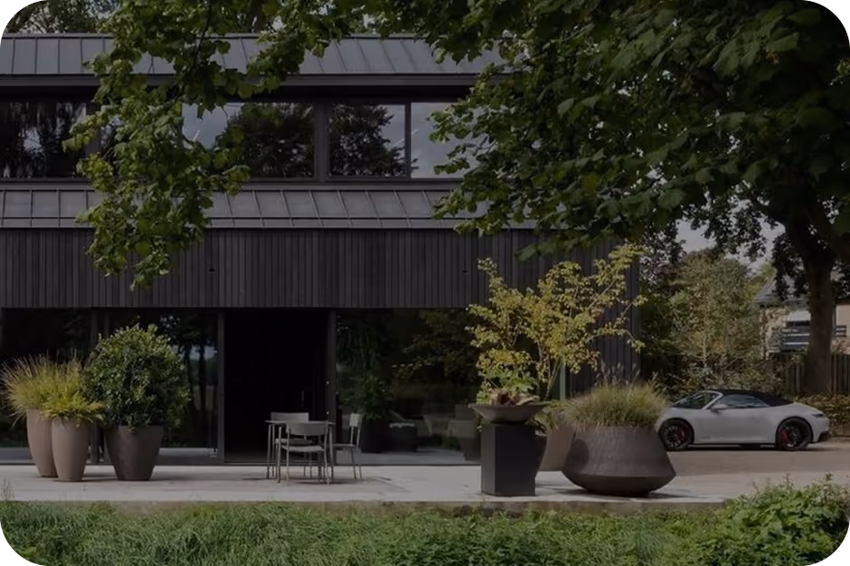 Modern black building with large windows, outdoor chairs, potted plants, and a white sports car parked nearby.