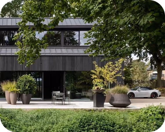 Modern house with large windows, outdoor chairs, potted plants, and a silver sports car parked nearby.